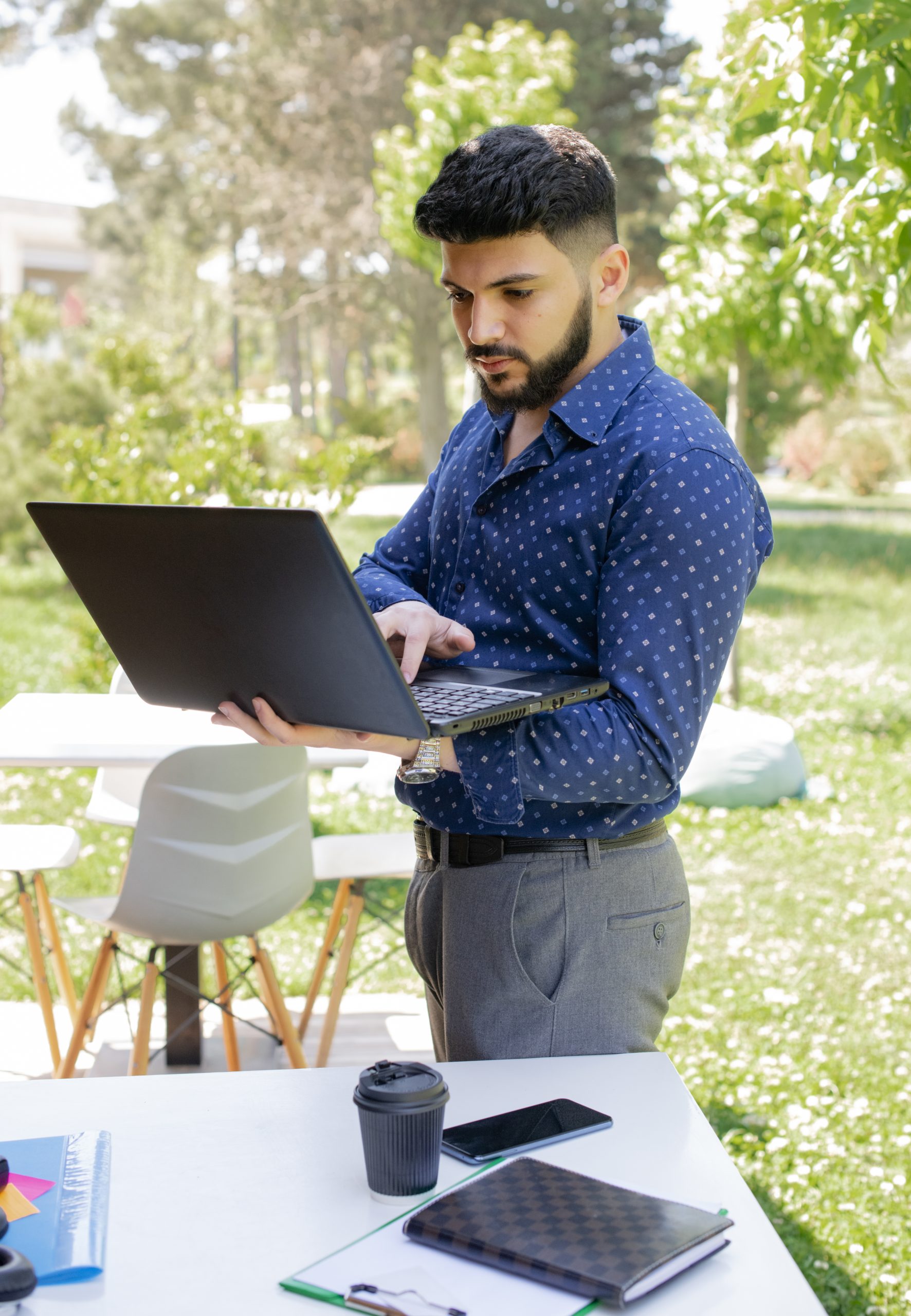 Handsome caucasian businessman typing on laptop outdoors. Portrait view of young black haired man in trendy shirt holding notebook, while working, with sunlit backyard on background. Concept of work.