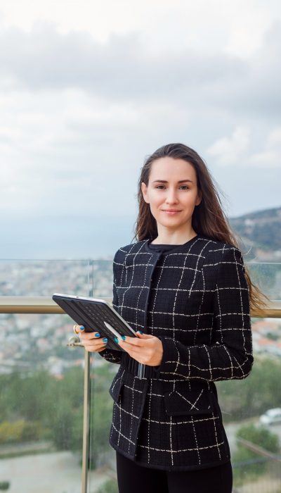 Blogger girl is looking at camera by holding planshet computer against the background of city view.